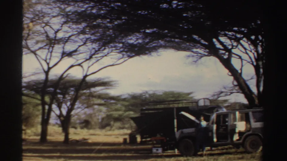 1969 Sudan home-movie frame showing a parked jeep with open doors beside a shelter under a broad tree in dry savanna.