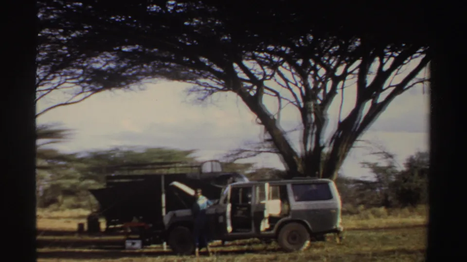 1969 Sudan home-movie frame with a person beside an open jeep near a simple camp shelter in a dry, tree-dotted landscape.
