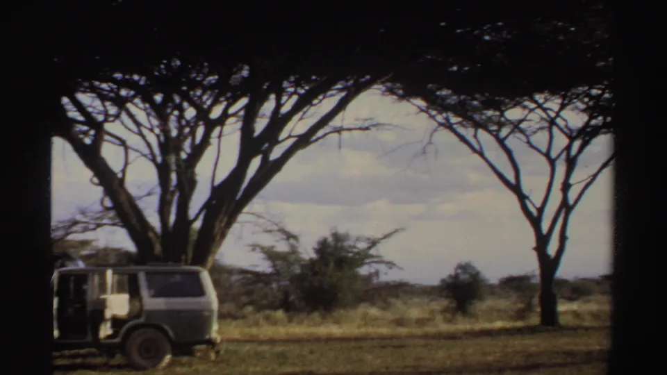 1969 Sudan 8mm frame with a parked vehicle under acacia-like trees, an open door, dry grassland, and cloudy sky.