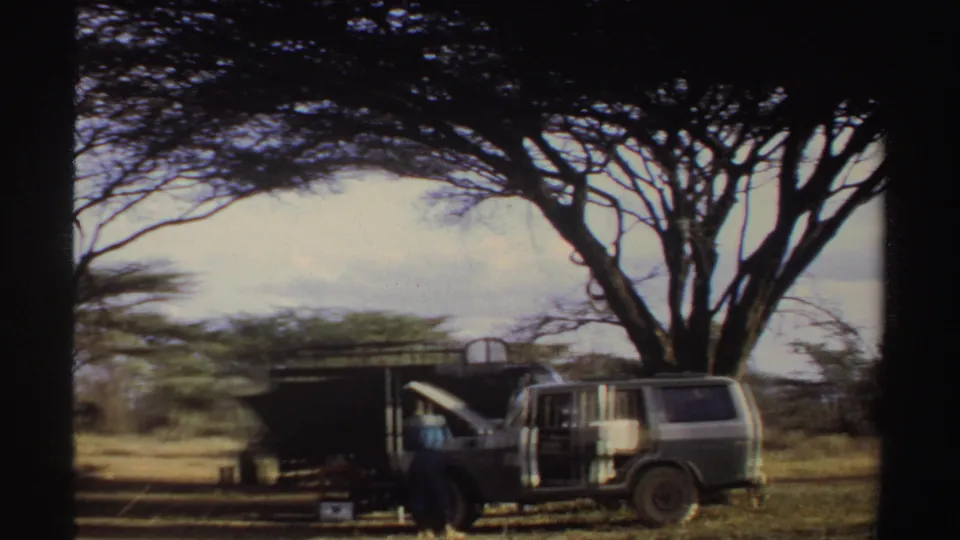 Sudan 1969 home-movie frame showing a parked vehicle near a dark shelter or equipment setup beneath a spreading acacia tree.