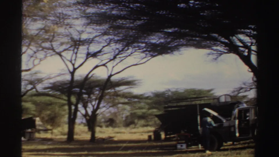 Sudan 1969 frame with acacia trees, a dark field structure, containers, and the edge of a parked vehicle in open savanna.