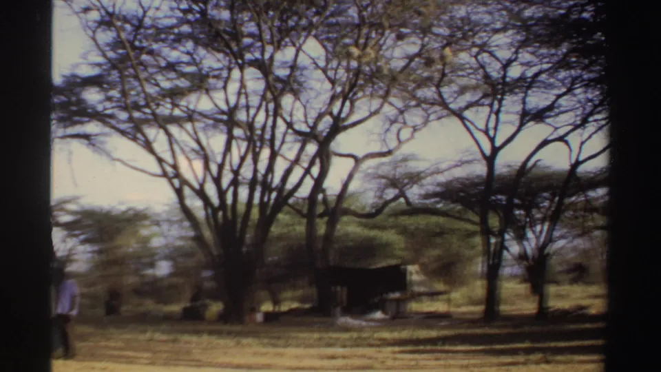 1969 Sudan 8mm frame with a person near a shaded camp shelter under sparse trees on dry ground.