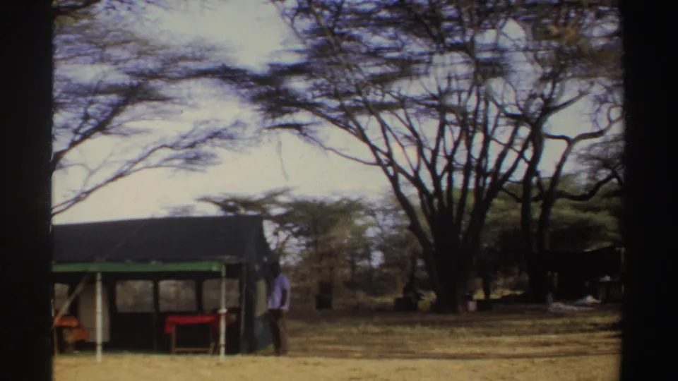 1969 Sudan 8mm frame showing a canvas tent with green trim, camp furniture, trees, and a nearby person.