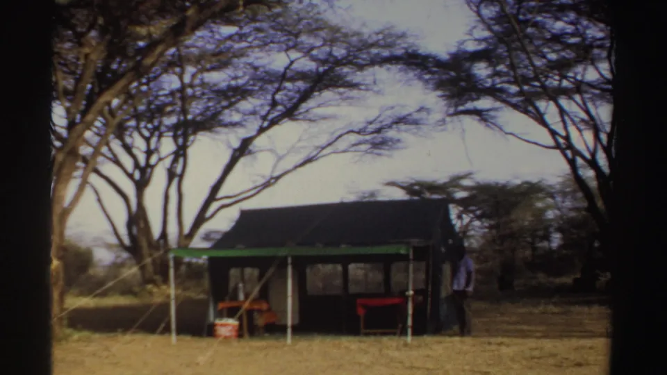1969 Sudan 8mm frame of a dark canvas tent with green trim, furniture, and a person beside it under large trees.