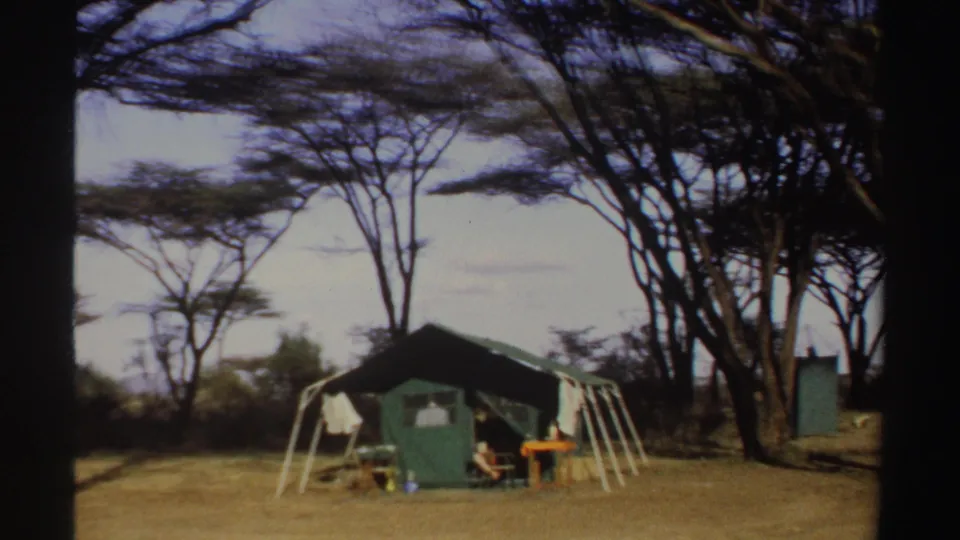 1969 Sudan 8mm frame showing a green tent, camp bed, small table, and portable toilet among sparse trees on dry ground.