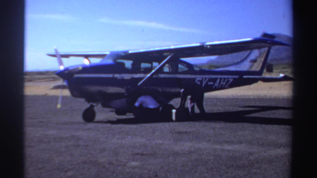 Small blue-and-white aircraft on a dusty airfield with a person crouched near luggage.
