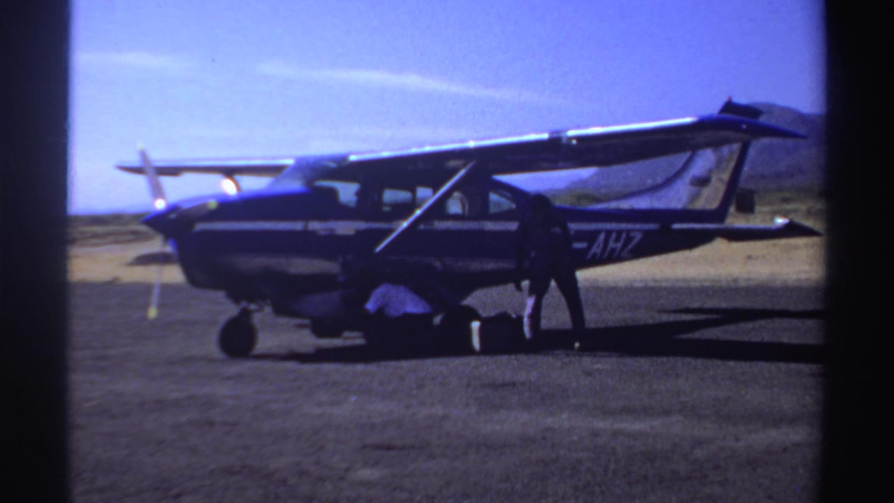 1969 Sudan frame of a small high-wing aircraft on a sunlit airfield as people work beside luggage near the fuselage.