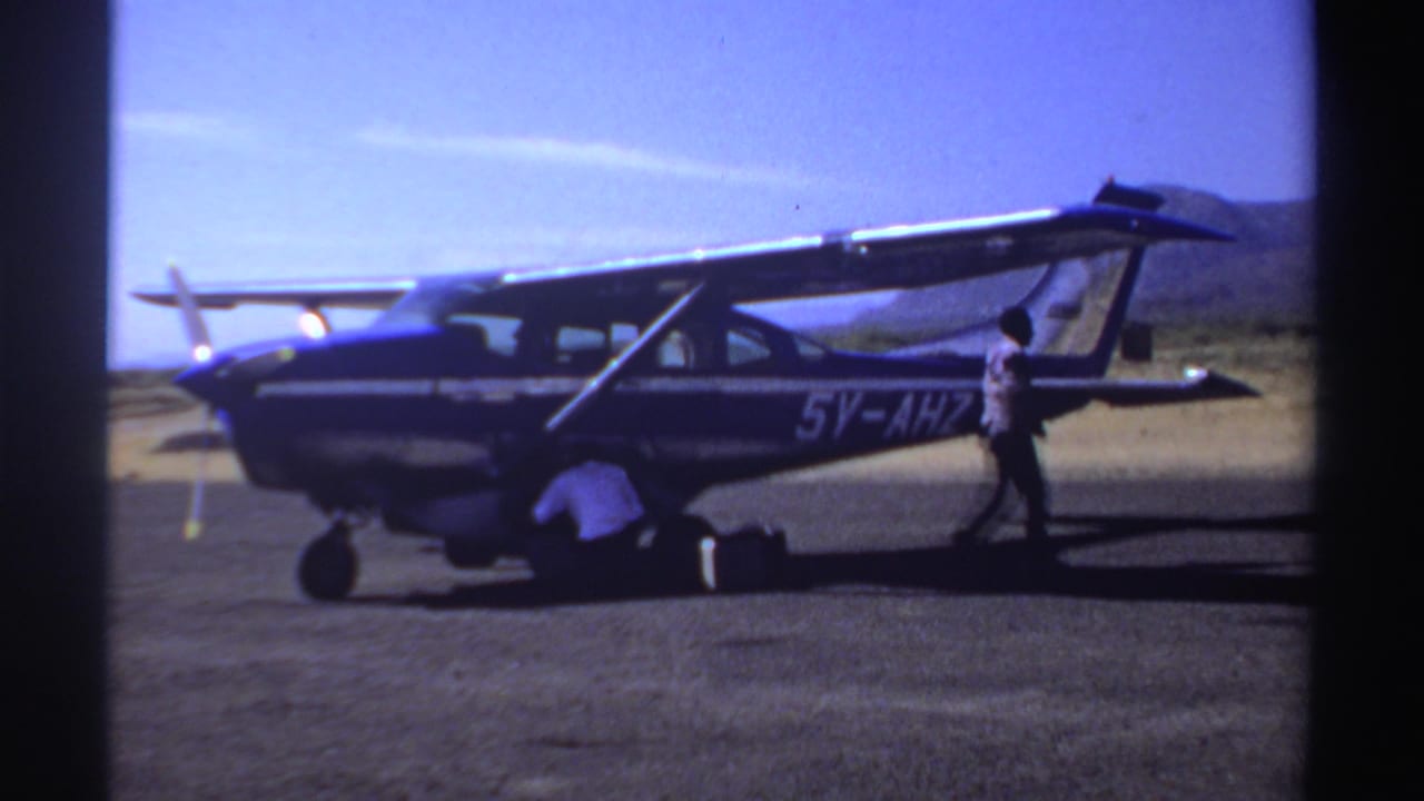 1969 Sudan home-movie frame showing the 5V-AHZ aircraft while one adult handles luggage and another stands near the rear.
