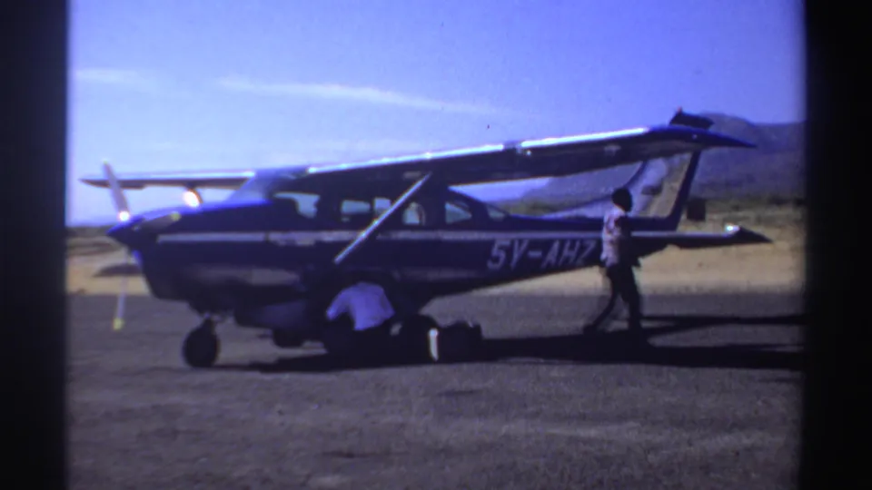 1969 Sudan home-movie frame showing the 5V-AHZ aircraft while one adult handles luggage and another stands near the rear.