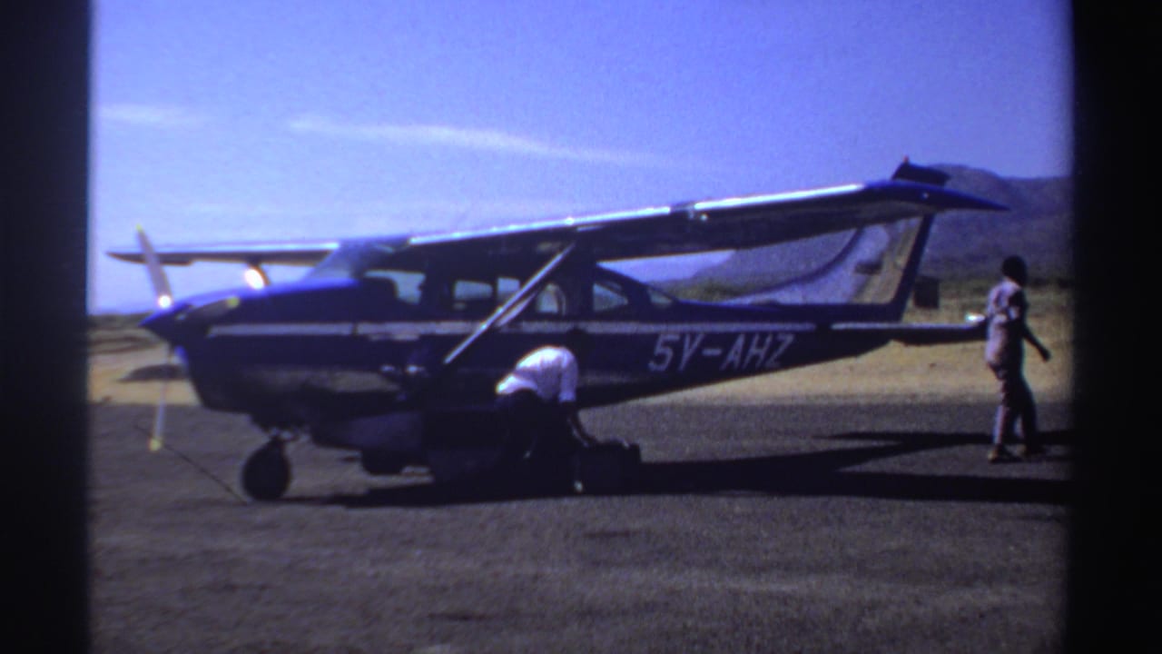 1969 Sudan frame with a person walking past the tail of aircraft 5V-AHZ as another person handles luggage near the cabin.