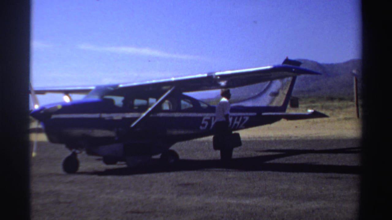 1969 Sudan frame with a man in a white shirt standing beside a parked blue and white airplane on an open airstrip.