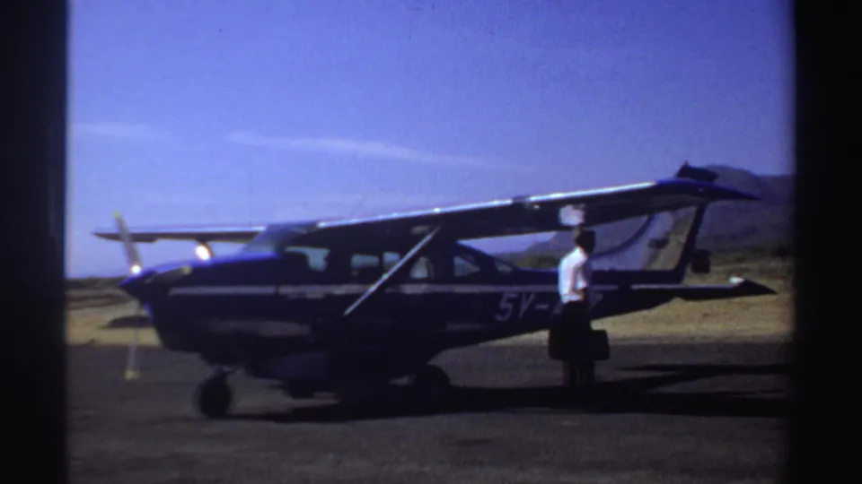 1969 Sudan home-movie frame showing a man with dark luggage beside the parked high-wing airplane on a bright airstrip.