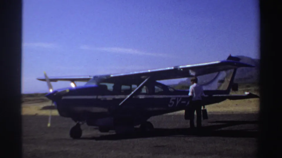 1969 Sudan frame showing a traveler with luggage beside a blue and white small airplane, with arid hills beyond the runway.