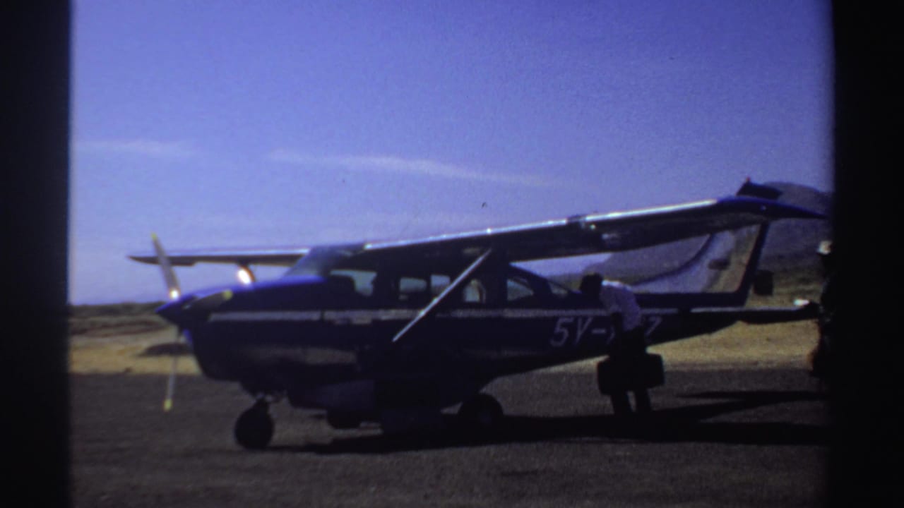 Blue and white high-wing aircraft on a dusty airstrip with people near the cabin.