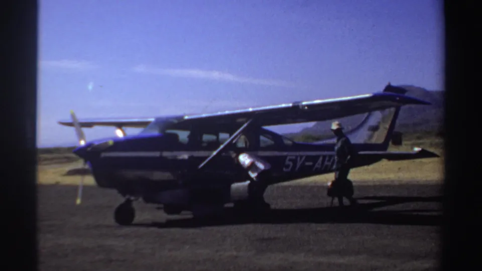 A person in a hat passes the open side of the blue and white aircraft on a sunny Sudan airstrip in 1969 footage.