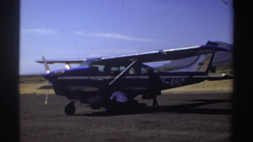 1969 Sudan frame with a man in a white shirt bending to handle luggage beside the marked blue and white aircraft.