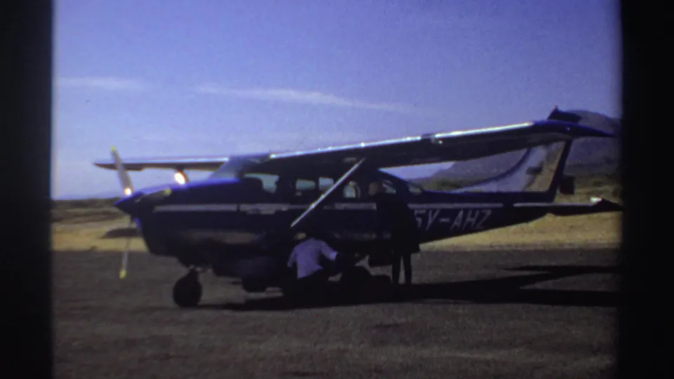 A man crouches near baggage at the open side of the blue and white aircraft, with another person standing nearby in 1969 Sudan footage.