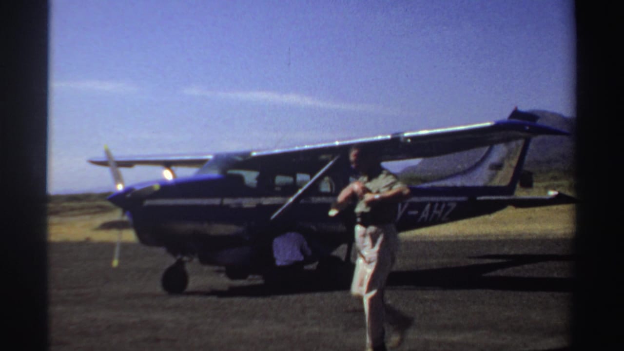 Man walks in front of a parked light aircraft while another person crouches by the door.
