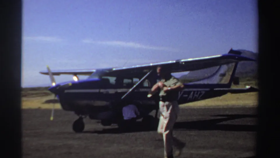 1969 Sudan home-movie frame with a man crossing the tarmac before a parked light aircraft as luggage is handled near the open door.