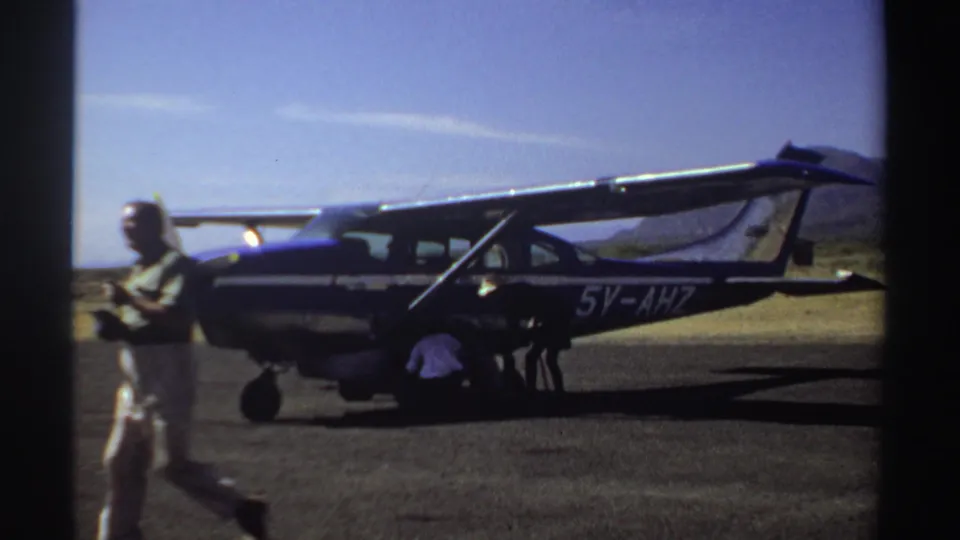 1969 Sudan frame showing a man in the foreground and people beside a small aircraft marked 5Y-AHZ on a bright airfield.