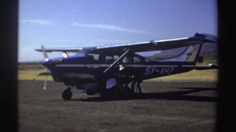 1969 Sudan 8mm frame of a parked single-propeller aircraft with people low beside the open cabin door on the airfield.