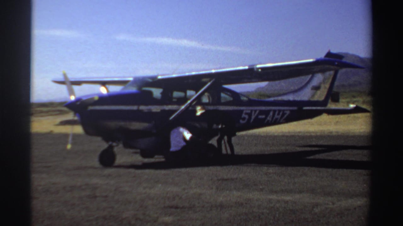 Small blue and white plane on a dusty airstrip with a man near the engine.