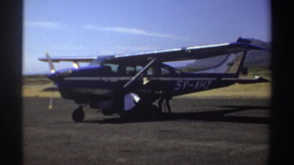 1969 Sudan home-movie frame showing a small blue and white aircraft marked 5V-AHZ as a man works near the engine.