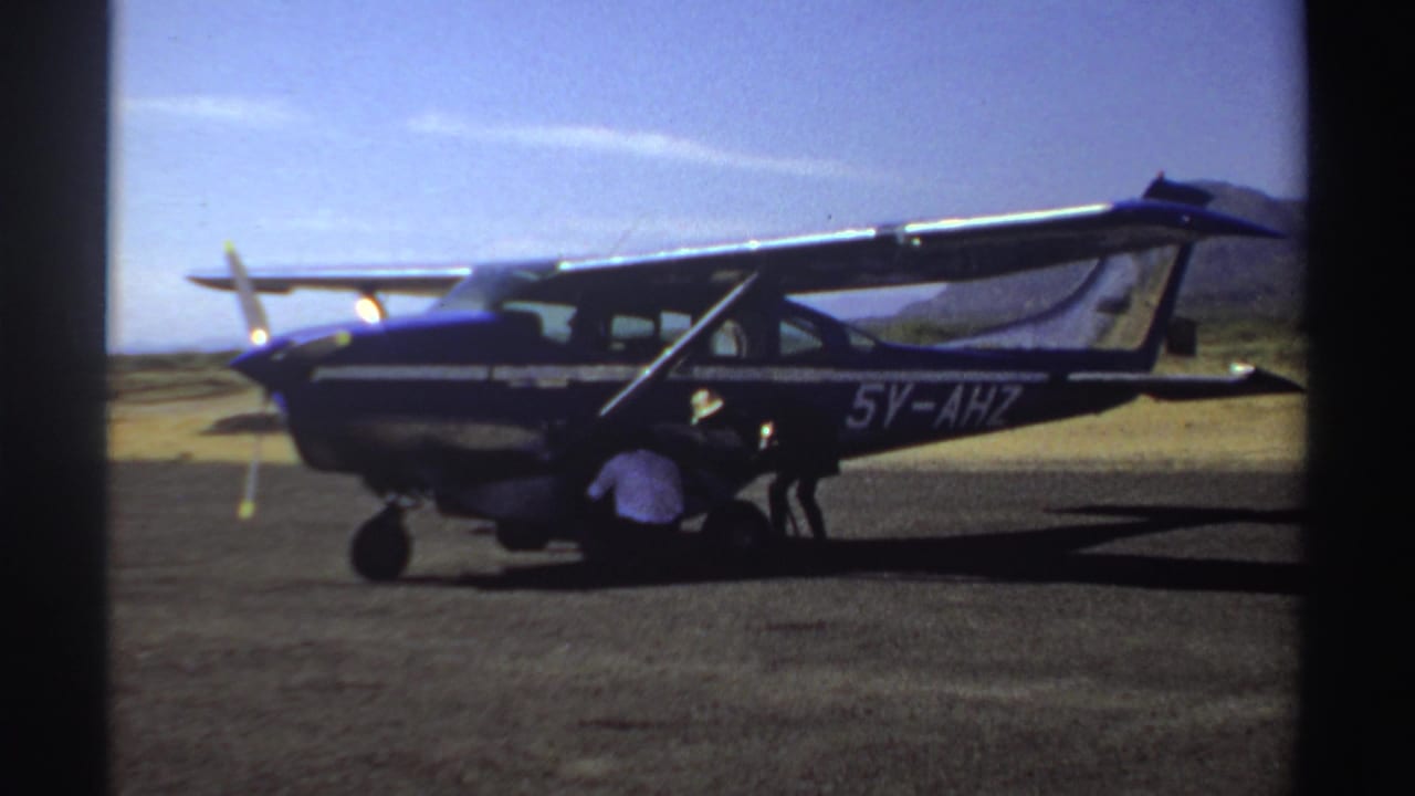 1969 Sudan frame of a blue and white single-engine plane on an airstrip while a man crouches near the front cowling.