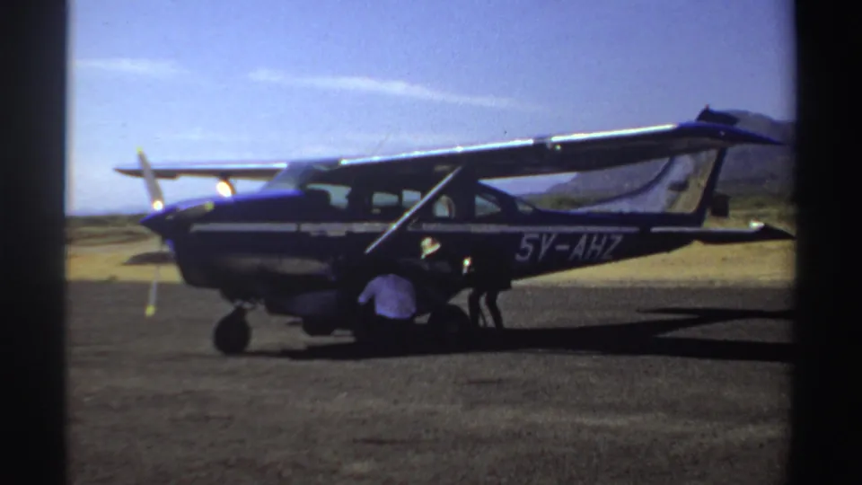 1969 Sudan frame of a blue and white single-engine plane on an airstrip while a man crouches near the front cowling.