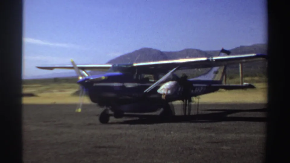 1969 Sudan home-movie frame showing men near a parked light aircraft with arid hills or mountains in the background.