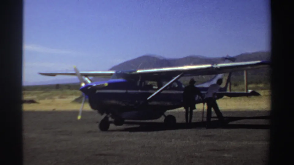 1969 Sudan frame of two men beside a blue and white light aircraft on a remote-looking airstrip with mountains behind.