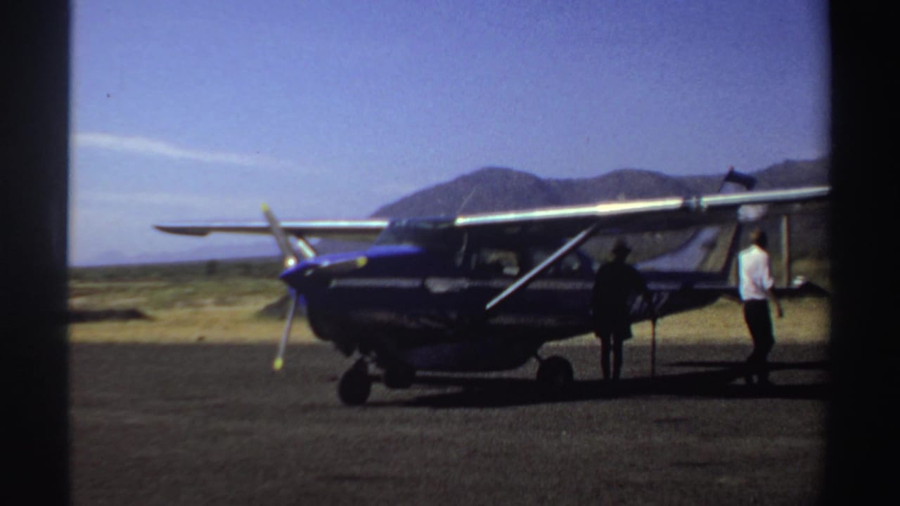 Blue and white single-engine plane parked on a dusty airstrip with two people nearby.