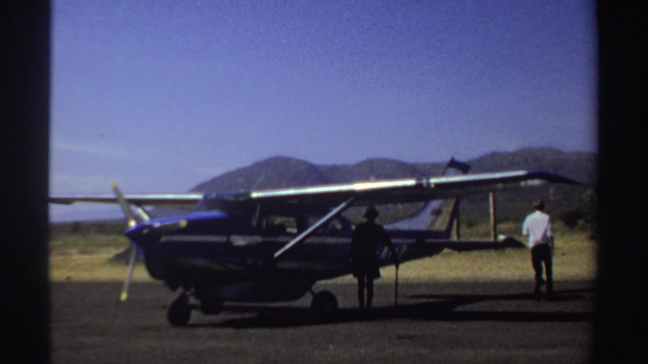 Parked single-engine plane with one figure by the fuselage and another near the tail.