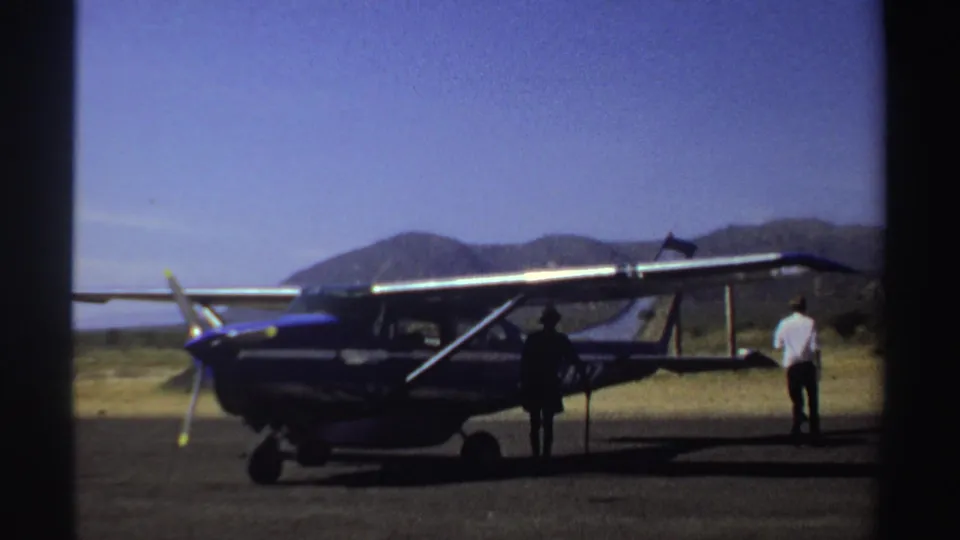 1969 Sudan frame with a small blue and white aircraft parked on an open airstrip, with two adults standing near it.