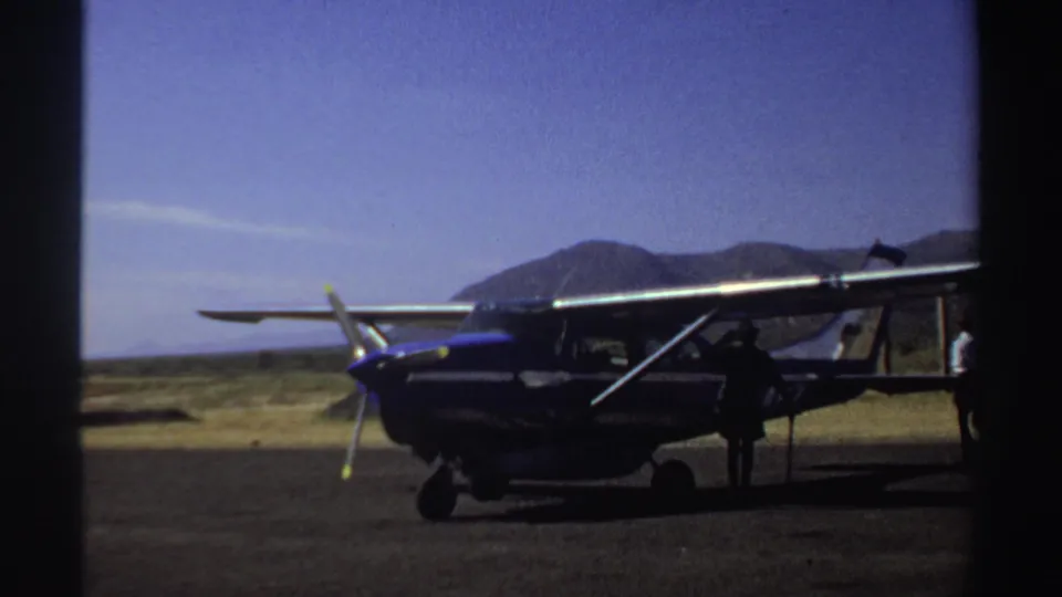 Sudan 1969 home-movie still showing a blue and white plane on a dusty airstrip, with a figure silhouetted beside it.