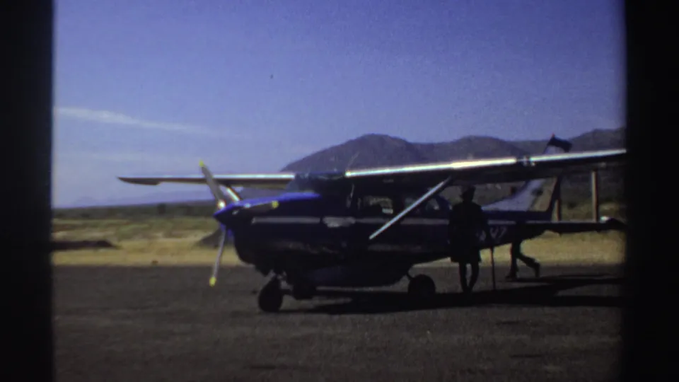 1969 Sudan footage frame of a small plane parked against distant hills, with people moving around the aircraft.