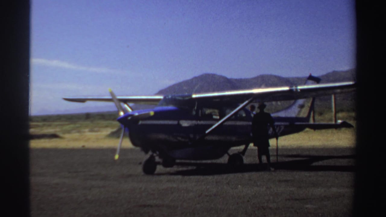 Blue-and-white single-engine plane parked on a dusty airstrip with a person standing near the cabin.