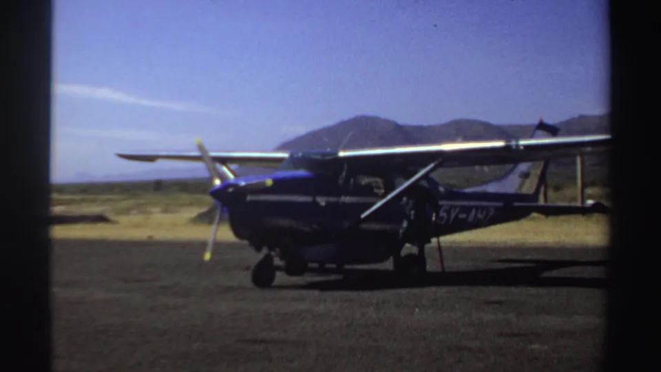 1969 Sudan 8mm still showing a parked charter plane with visible side markings, landing gear, dry ground, and mountains beyond.