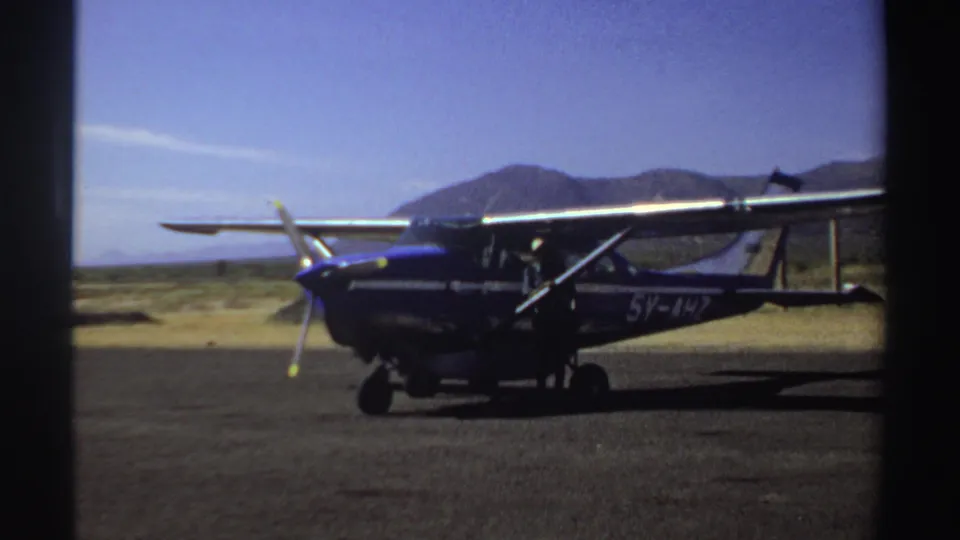 Late-1960s Sudan 8mm frame of a parked single-engine plane in profile, seen against open sky and a distant mountain ridge.
