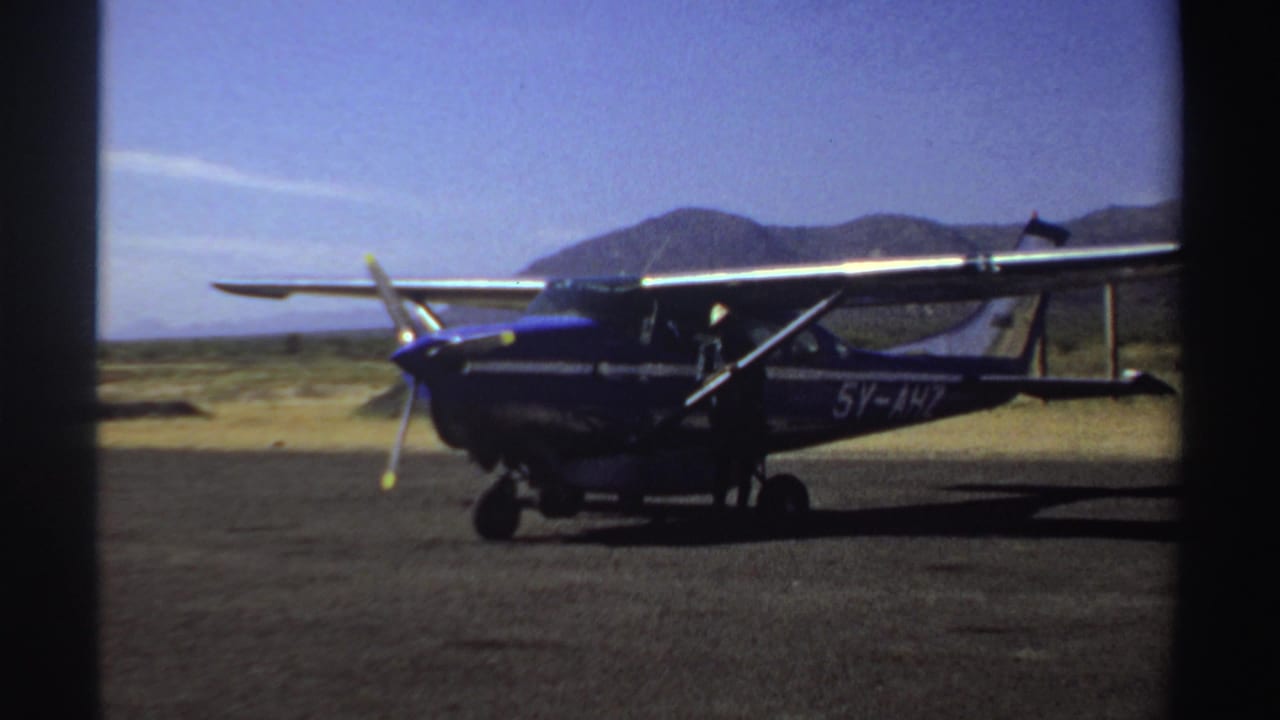 Side view of the blue-and-white light aircraft at a Sudan airstrip in 1969, framed by open terrain and mountains.