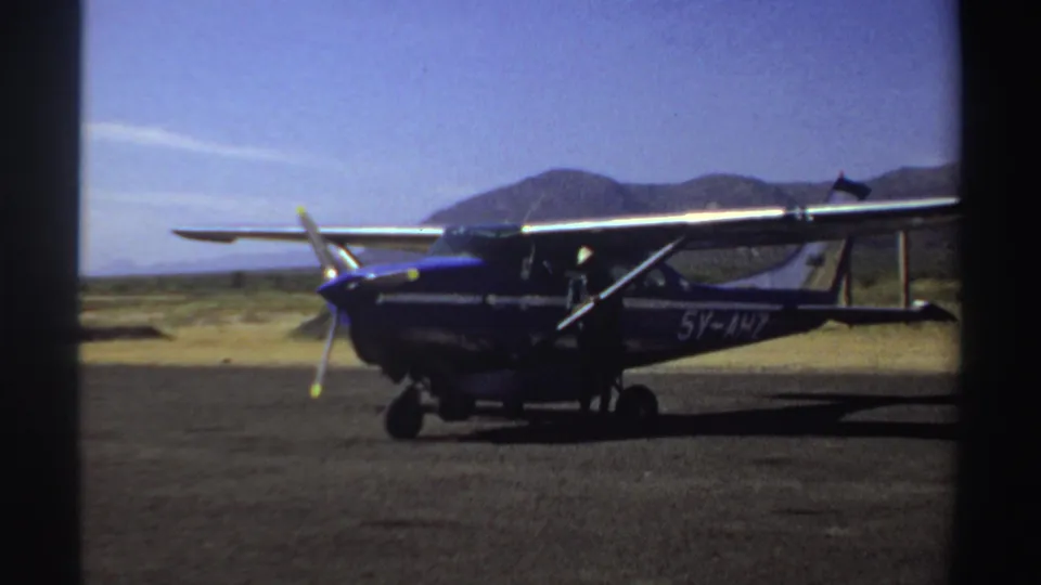 Side view of the blue-and-white light aircraft at a Sudan airstrip in 1969, framed by open terrain and mountains.