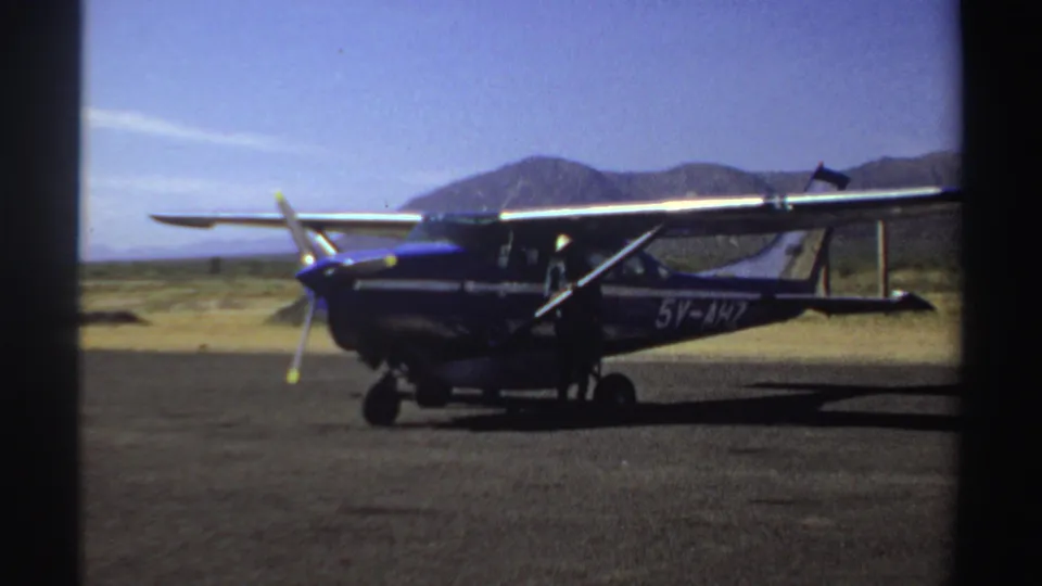 The 1969 Sudan home-movie frame holds on the aircraft parked at the remote strip, with dry ground and hills behind it.