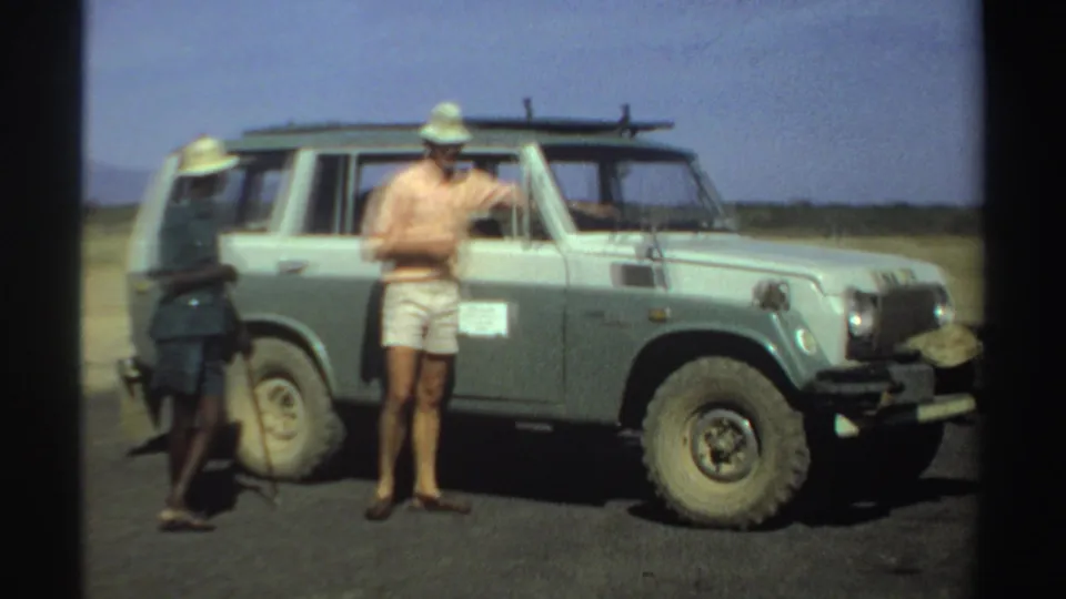 Two men at a Sudan airstrip in 1969 stand by a light-colored jeep, one in shorts gesturing and another in a dark uniform and hat.