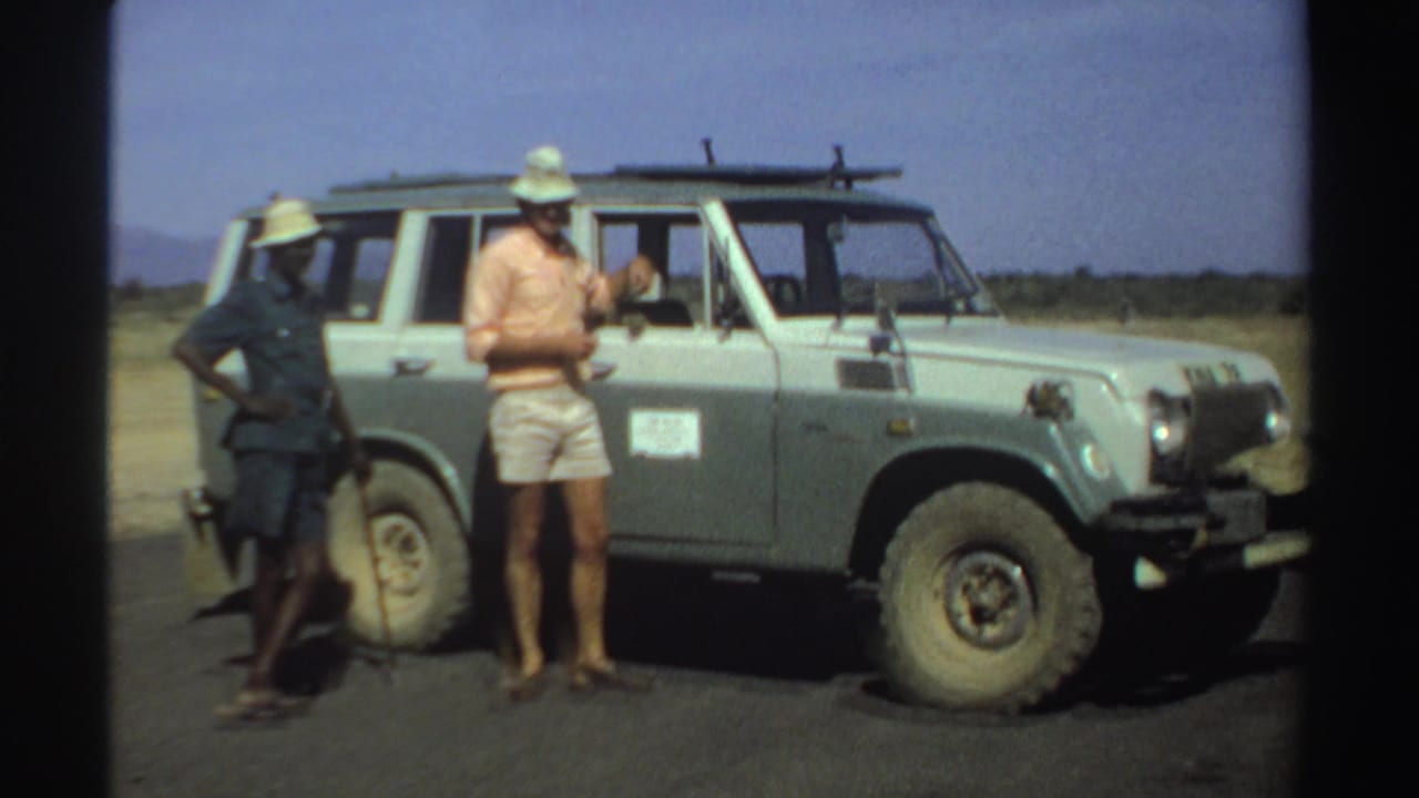 Two adults stand beside a light off-road vehicle on a dry roadside.