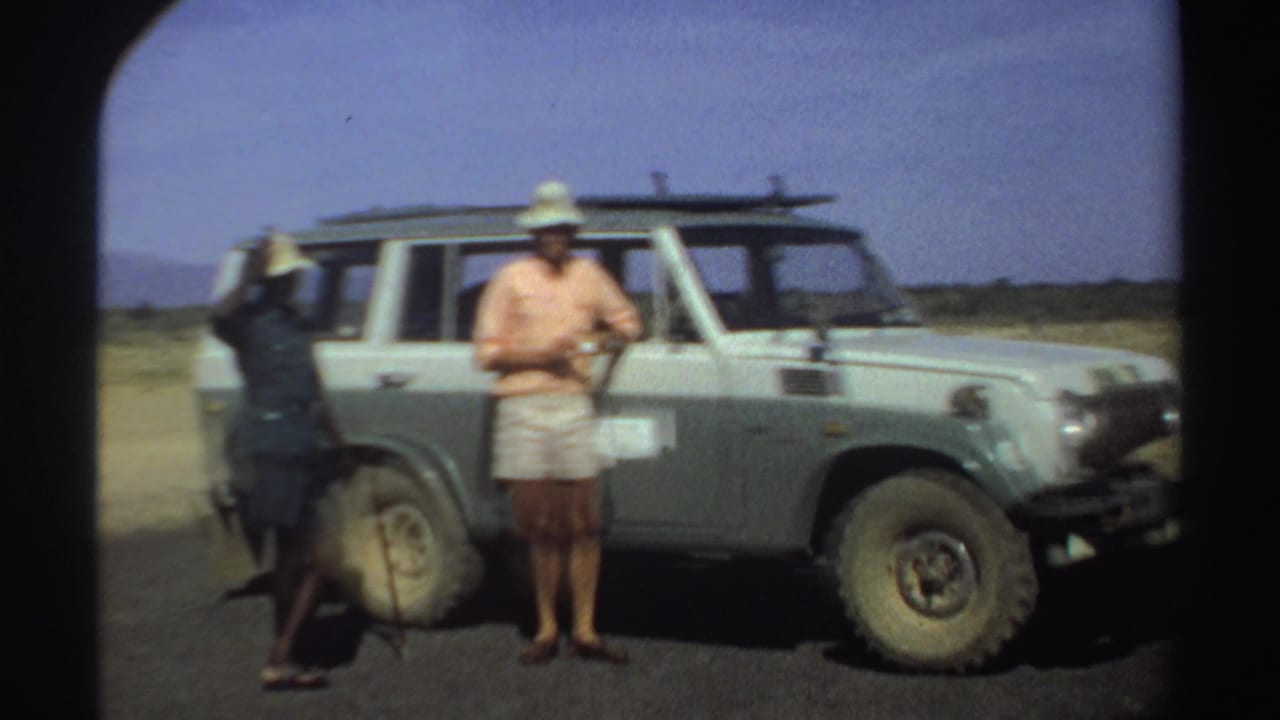 1969 Sudan travel footage frame with a man in a pink shirt standing by a light off-road vehicle and another adult nearby.