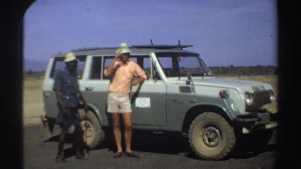 1969 Sudan home-movie frame showing a man adjusting his hat beside a light off-road vehicle on open terrain.