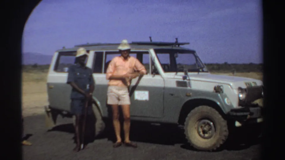 1969 Sudan frame with two adults standing near a parked light off-road vehicle under a clear sky.
