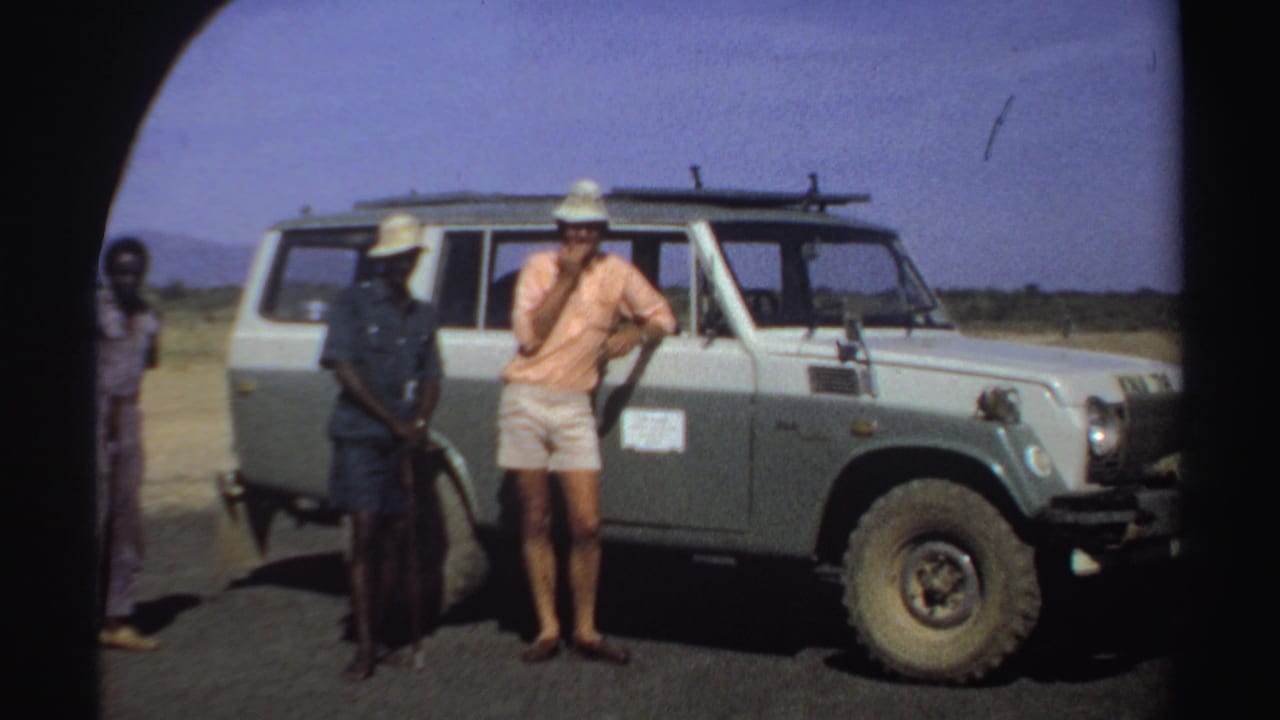 Three men stand beside a light-colored safari vehicle on a sunny road.
