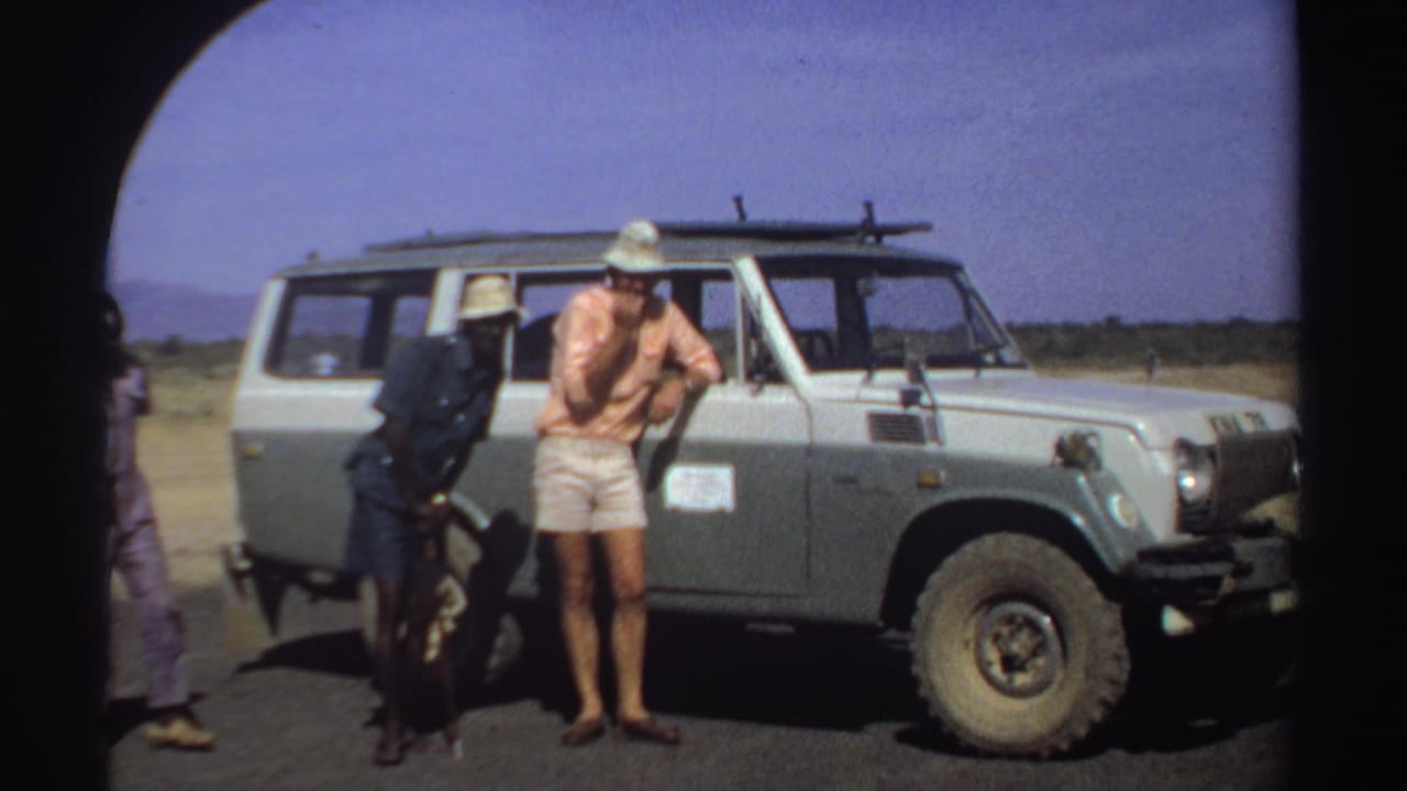 Men in sun hats stand near the side and front of a safari vehicle.