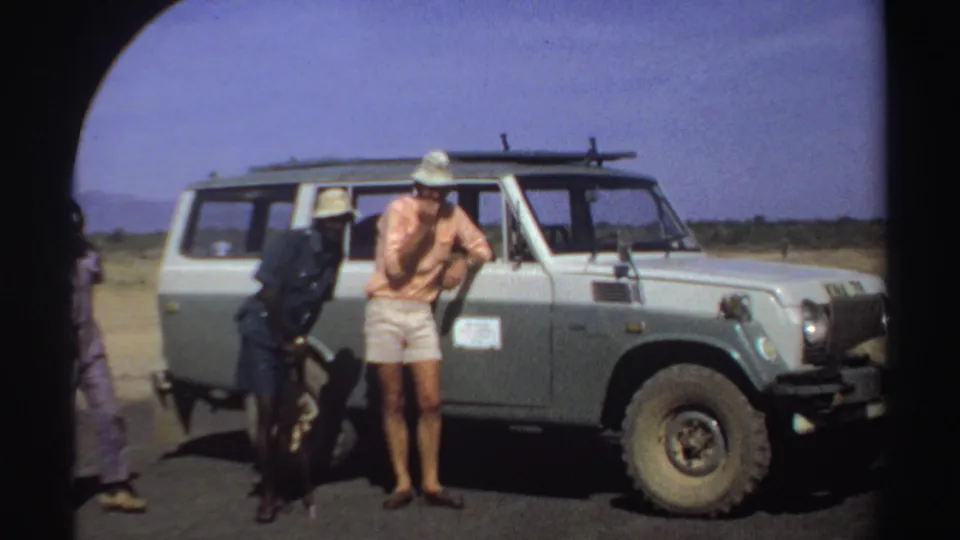 Three men in sun hats appear beside a vintage off-road vehicle in a bright Sudan roadside scene from 1969.
